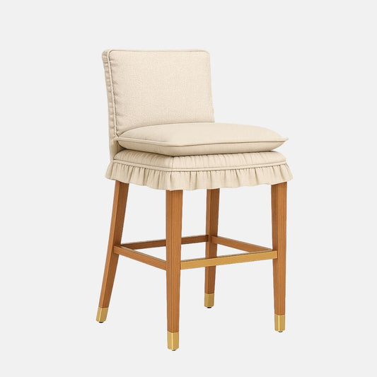 Cream upholstered bar stool with a ruffled skirted seat, square backrest, wooden legs with brass caps and a brass footrest, on a white background