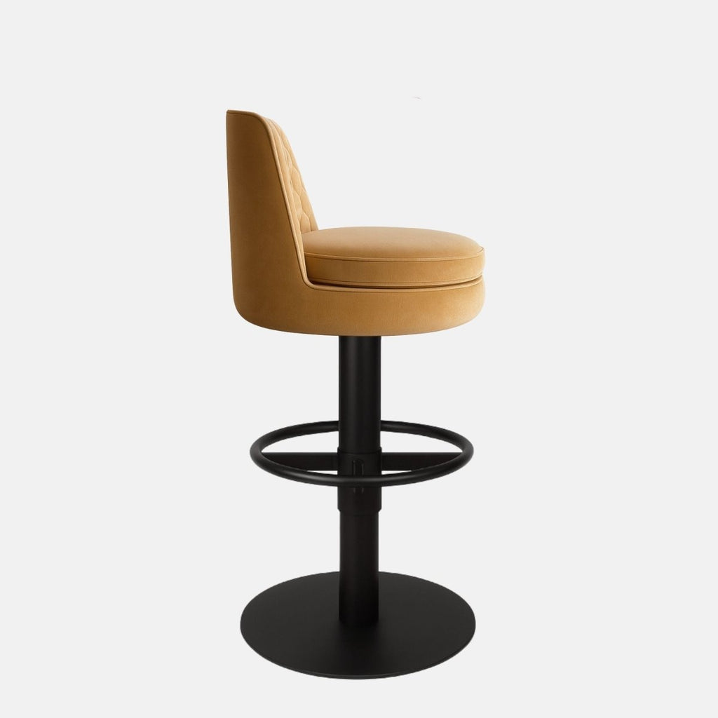 Mustard yellow upholstered bar stool with a hexagonal quilted backrest, round cushioned seat, black circular footrest, and round black metal base on a white background