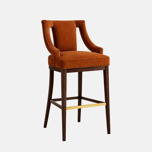 Front three-quarter view of a burnt-orange velvet bar stool with an open-back design and slim upholstered arms, on dark wood legs with a brass footrest bar, photographed on a white background.