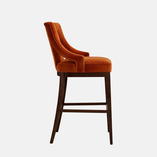 Side profile of a burnt-orange velvet bar stool with a tall, gently curved back and slim upholstered arms, set on dark wood legs with a lower stretcher, photographed on a white background.