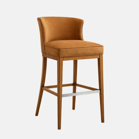 Caramel/tan upholstered bar-height stool with a curved low back and thick cushioned seat, on a warm oak wooden frame with four tapered legs, square stretchers, and a brushed metal front footrest, photographed on a white background.