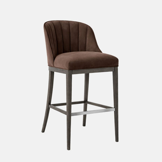 Three-quarter view of a bar-height stool with a brown upholstered seat and curved, channel-stitched backrest on a dark grey wooden frame with four legs and a brushed-metal footrest bar, photographed on a white background.