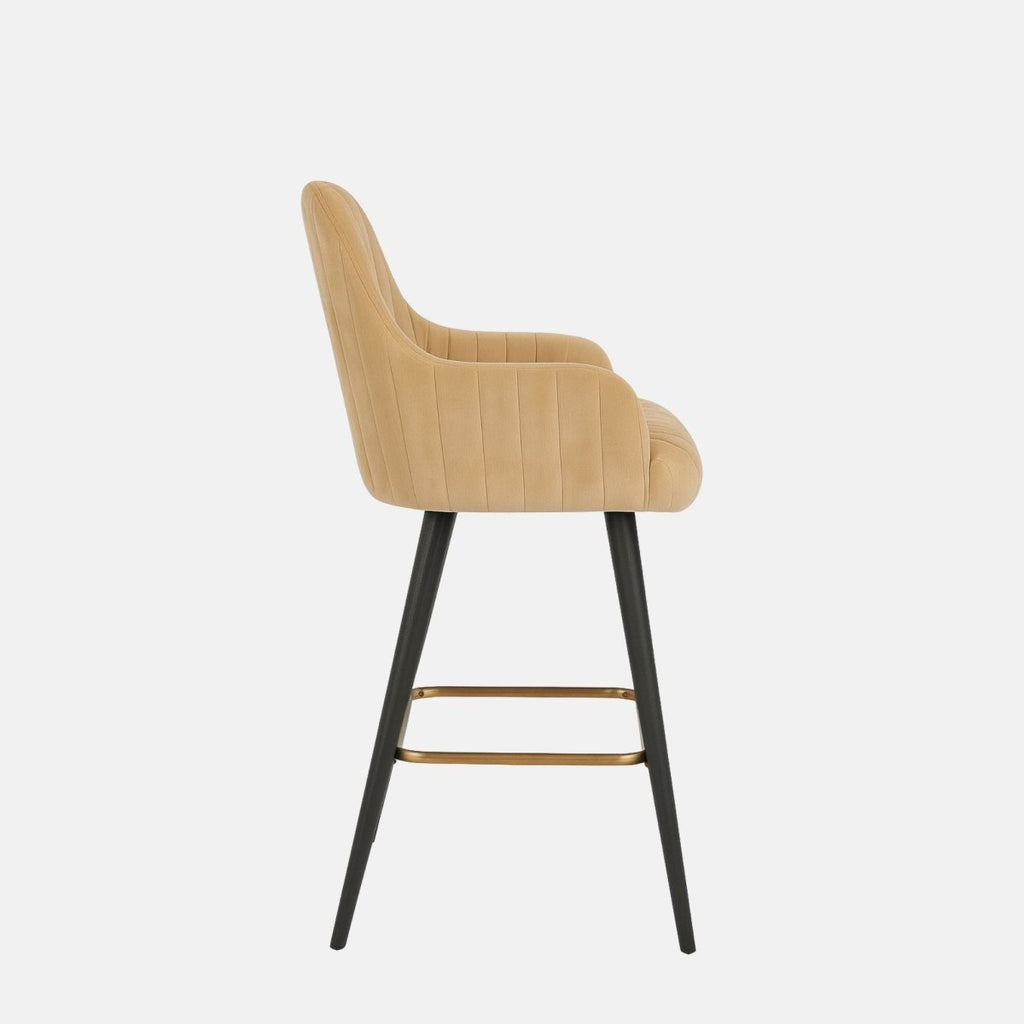 Beige upholstered bar stool with vertical channel stitching, curved armrests, black tapered legs, and a brass footrest.