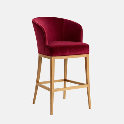 Three-quarter view of a bar-height stool with a curved, wraparound tub back and a thick seat cushion in deep raspberry/burgundy velvet (subtle ribbed texture on the seat), on a light wood four-leg frame with square stretchers, photographed against a white background