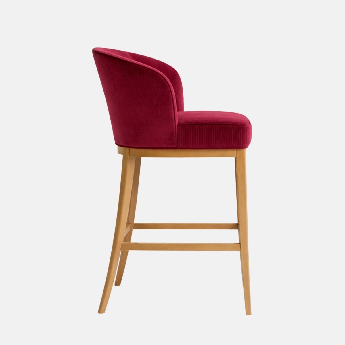 Side view of a bar-height stool with a rounded tub-style back and thick upholstered seat in deep raspberry/magenta velvet, on a light wood four-leg frame with two horizontal stretchers, photographed on a white background.