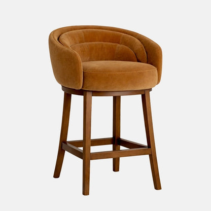 Front view of a caramel-brown velvet bar stool with a rounded tub-style back and curved arms, cushioned seat, and dark wooden legs with a square footrest frame, photographed on a white background.