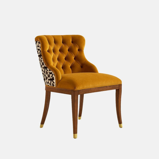 Upholstered accent chair in mustard-gold velvet with a button-tufted backrest, curved silhouette, patterned fabric on the outer back, and dark wooden legs with brass-capped feet, shown on a white background