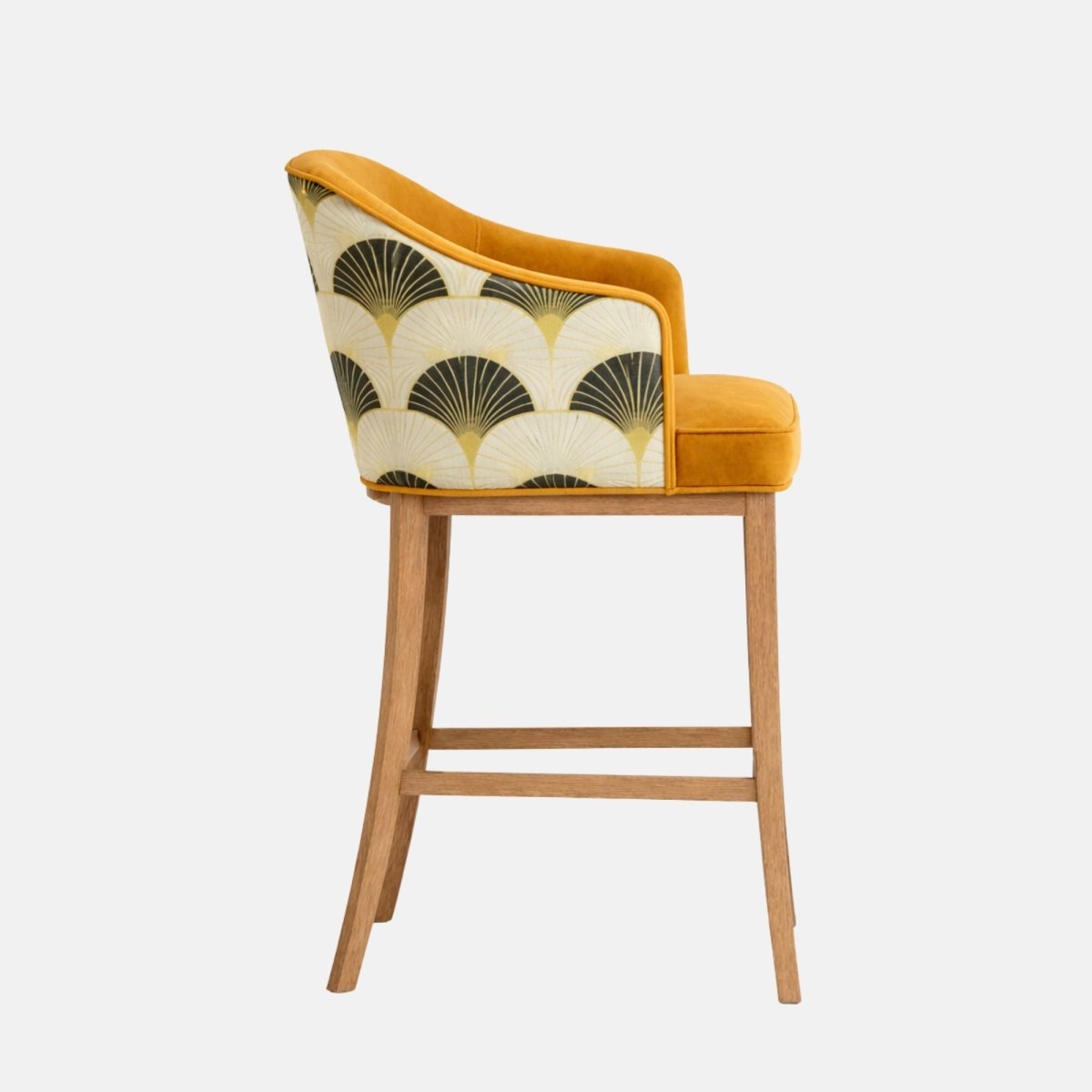 Side profile of a bar-height stool with a curved wraparound back and low arms, upholstered in mustard-gold velvet with an art-deco fan-pattern fabric on the outer back, on a light wood four-leg frame with horizontal stretchers, photographed on a white background.