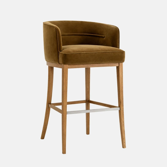 Bar-height upholstered stool in olive/mustard velvet with a curved wraparound back, thick cushioned seat, and a light oak wooden frame with square stretchers and a brushed metal footrest, photographed on a white background.