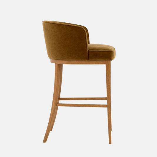 Side view of a bar-height stool upholstered in olive/mustard velvet with a curved wraparound back and thick cushioned seat, set on a light oak wooden frame with four legs and stretchers, photographed on a white background.