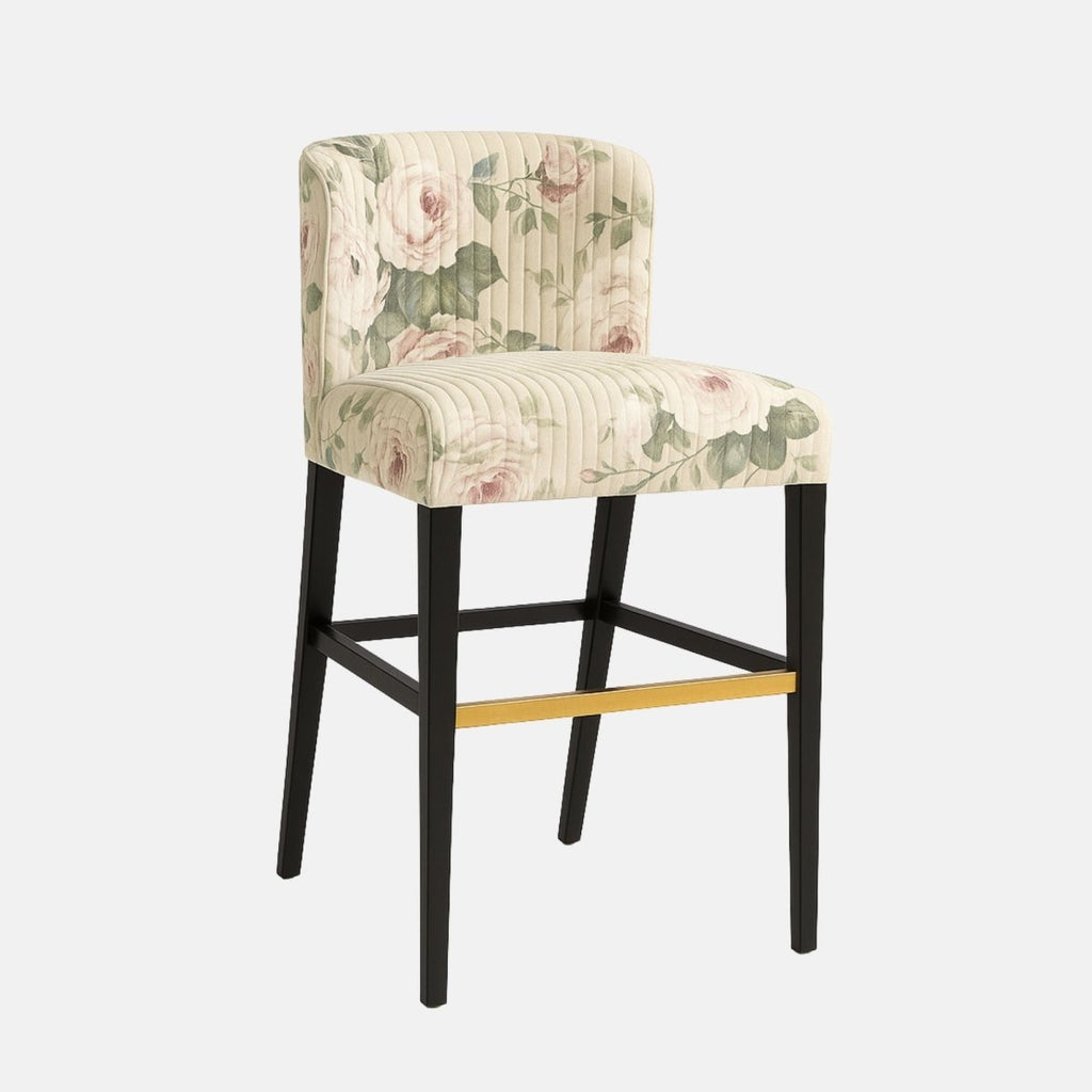 Elegant bar stool with a curved backrest and vertical stitching, upholstered in cream fabric featuring a soft floral pattern with pink roses and green leaves. It has a black wooden frame with a brass footrest accent, blending classic charm with modern style.