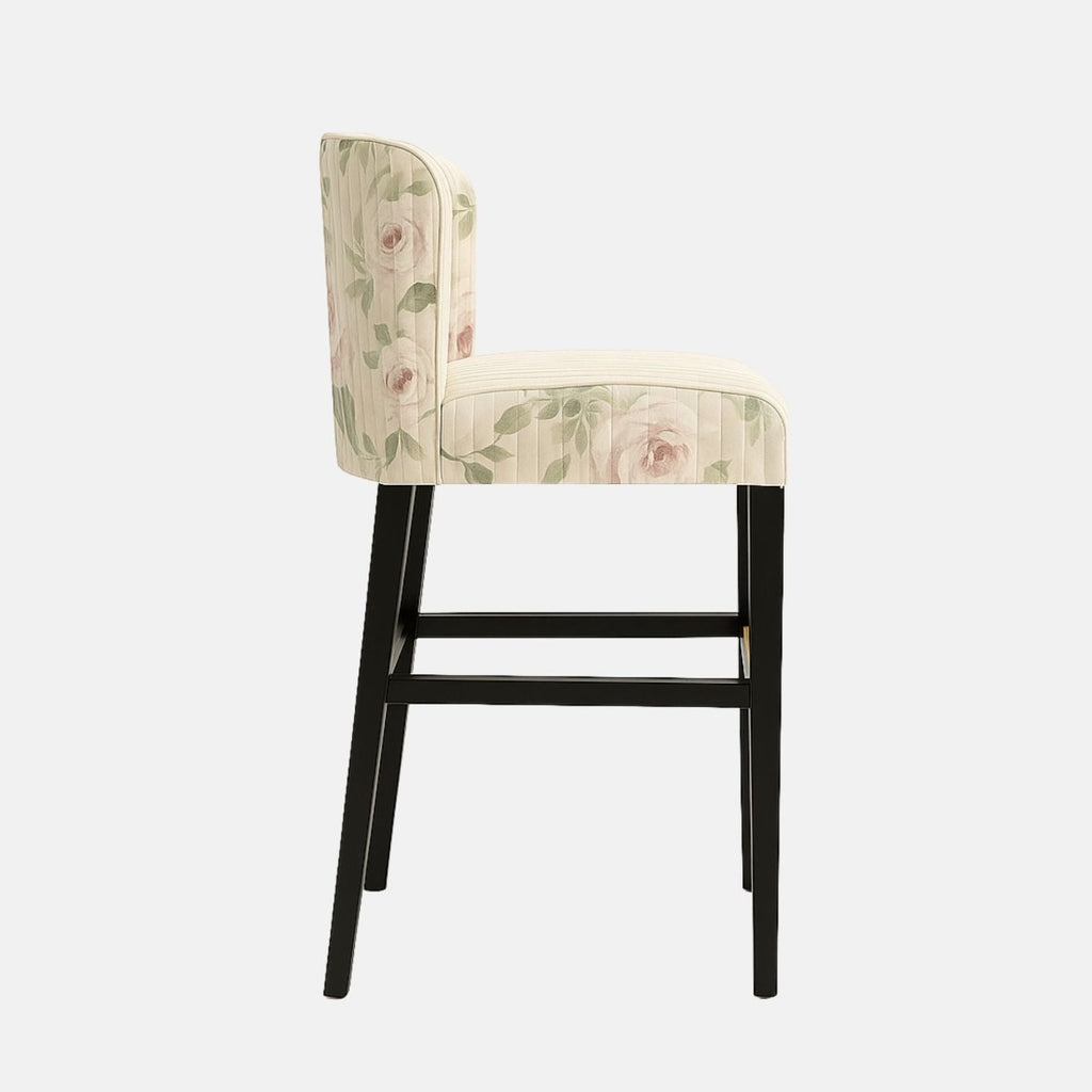 Elegant bar stool with a curved backrest and vertical stitching, upholstered in cream fabric featuring a soft floral pattern with pink roses and green leaves. It has a black wooden frame with a brass footrest accent, blending classic charm with modern style.