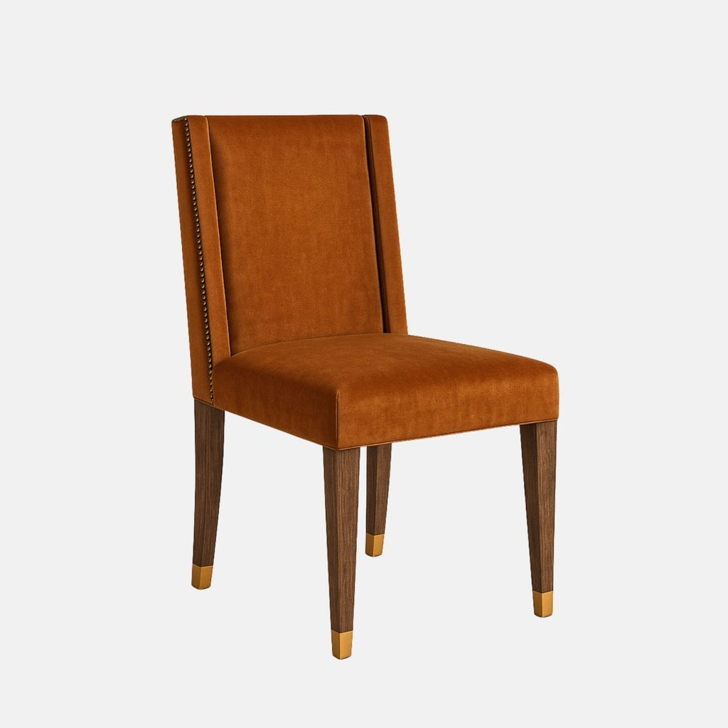 Upholstered dining chair in rich tan leather with a straight padded backrest, cushioned seat, nailhead trim along the back edges, and dark wooden legs with brass-capped feet, shown on a white background.
