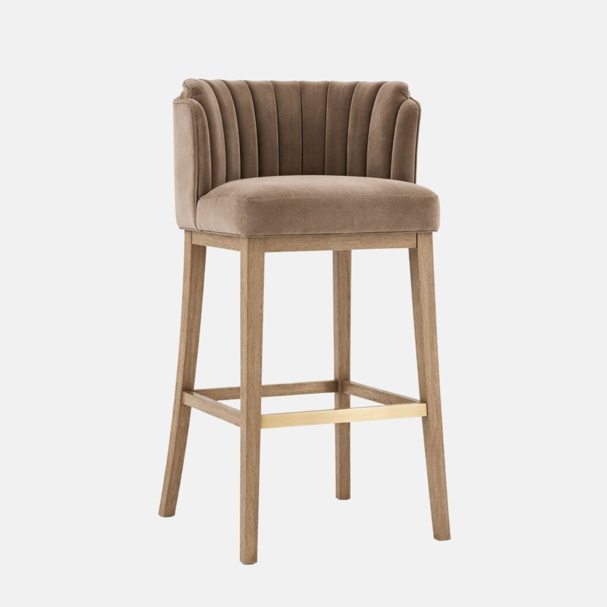 Three-quarter view of a taupe upholstered bar stool with a vertically channel-tufted, wraparound backrest and thick cushioned seat, on light oak wooden legs with a gold-toned footrest, photographed on a white background.
