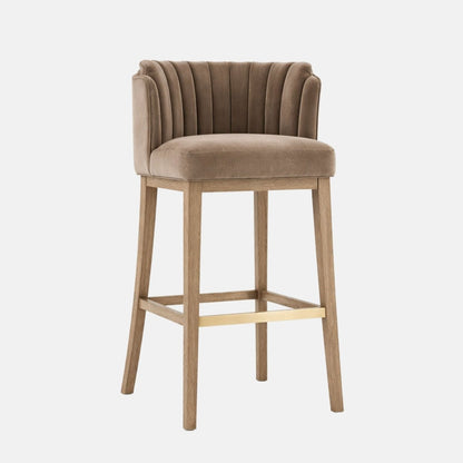 Three-quarter view of a taupe upholstered bar stool with a vertically channel-tufted, wraparound backrest and thick cushioned seat, on light oak wooden legs with a gold-toned footrest, photographed on a white background.