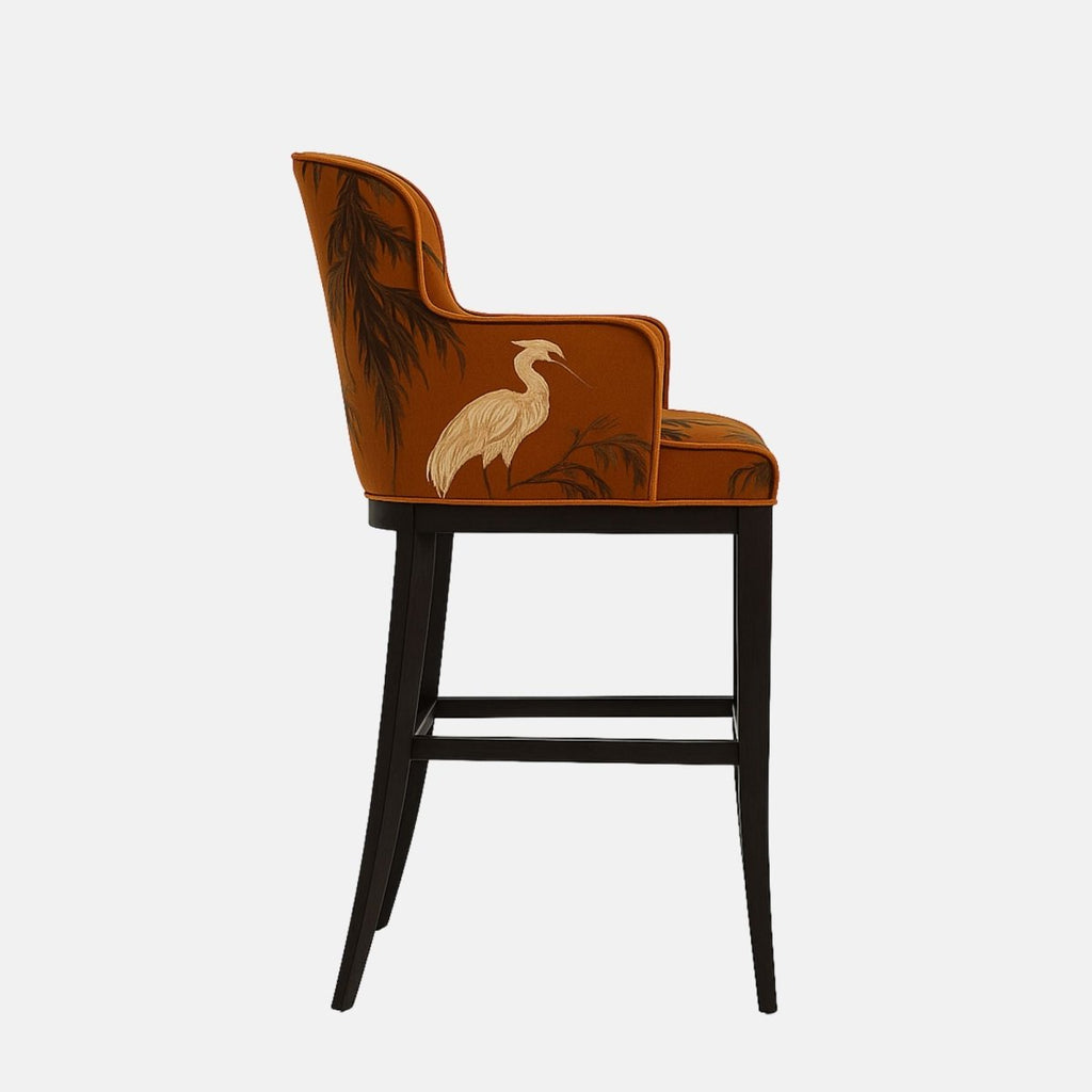 Elegant bar stool with a curved backrest and short armrests, upholstered in burnt orange fabric featuring a white heron and tree branch pattern. The seat is cushioned and supported by a black wooden frame with a brass footrest accent, blending artistic flair with classic design
