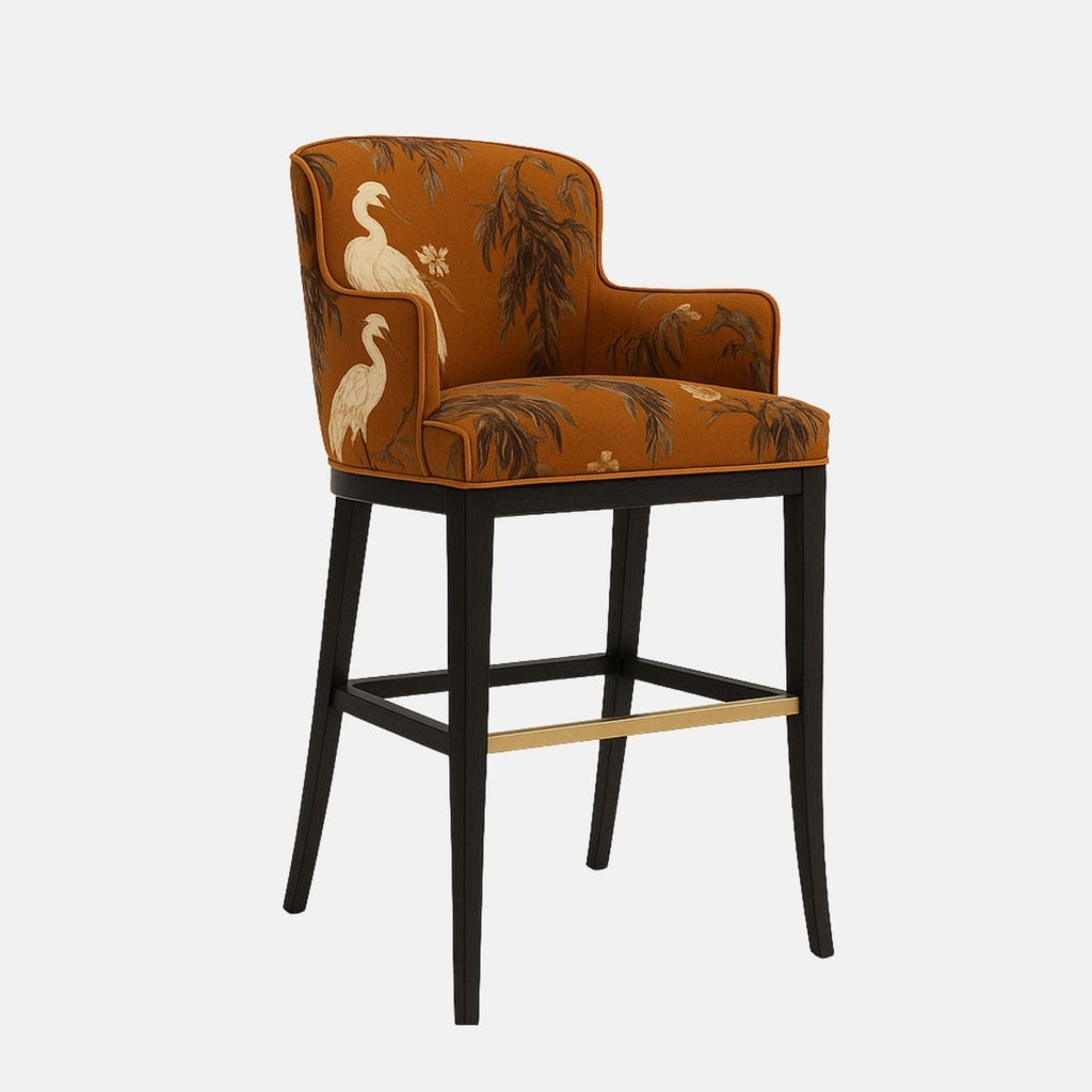 Elegant bar stool with a curved backrest and short armrests, upholstered in burnt orange fabric featuring a white heron and tree branch pattern. The seat is cushioned and supported by a black wooden frame with a brass footrest accent, blending artistic flair with classic design