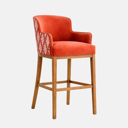 Three-quarter view of an orange upholstered bar stool with a curved wraparound back and integrated arms, a patterned fabric outer back panel, thick cushioned seat, and a light oak wooden frame with square stretchers, photographed on a white background.