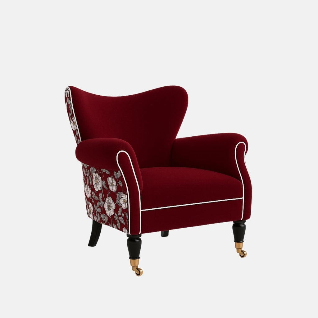 Deep red upholstered armchair with white piping, rolled arms, floral patterned fabric on the back and sides, black legs, and brass front castors.