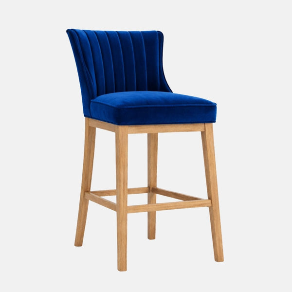 Royal blue velvet bar stool with a vertically channel-tufted backrest, padded seat, and light wood legs with a square stretcher/footrest.