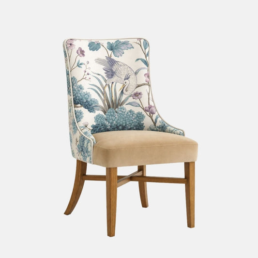 Upholstered dining chair with a beige cushioned seat and a high, gently curved backrest upholstered in a blue botanical and bird-patterned fabric, supported by natural wooden legs, shown on a white background