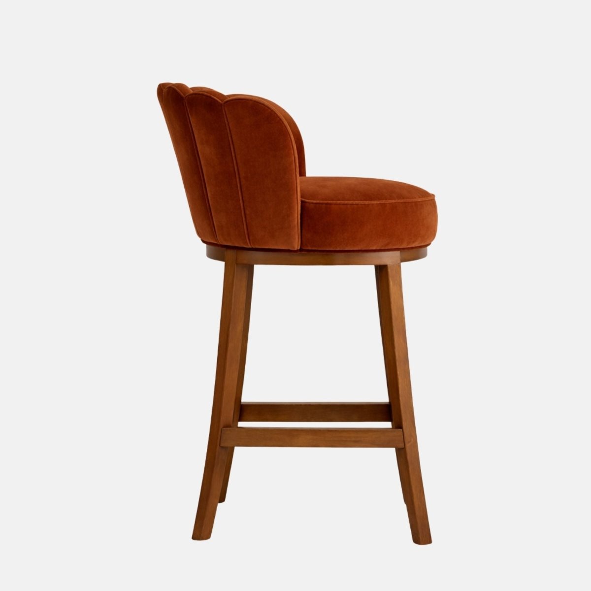 Side view of a burnt-orange velvet bar stool with a scalloped, channel-tufted back and a thick round seat, on angled medium-brown wooden legs with a rectangular footrest stretcher, photographed on a white background.