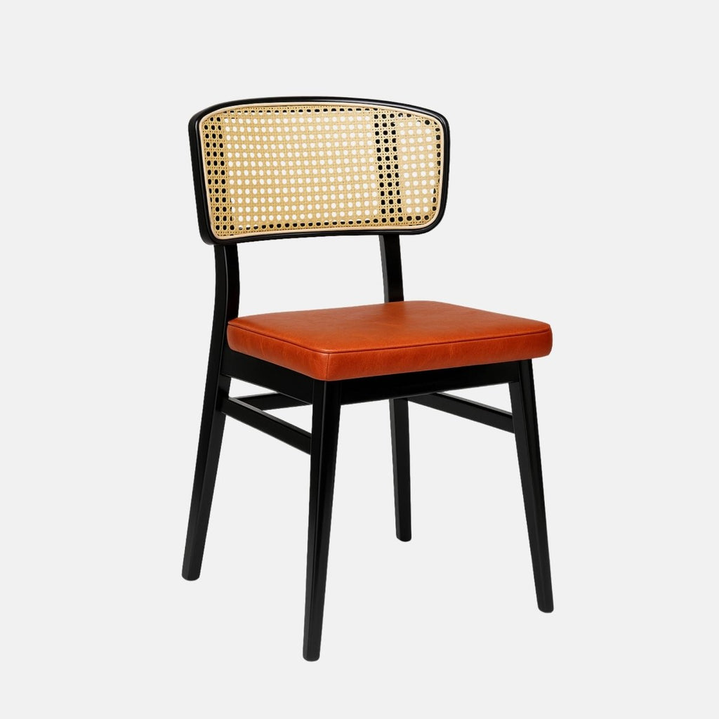 Contemporary dining chair with a black wooden frame, natural cane backrest, and a square padded seat upholstered in burnt orange leather. The design combines mid-century and modern styles