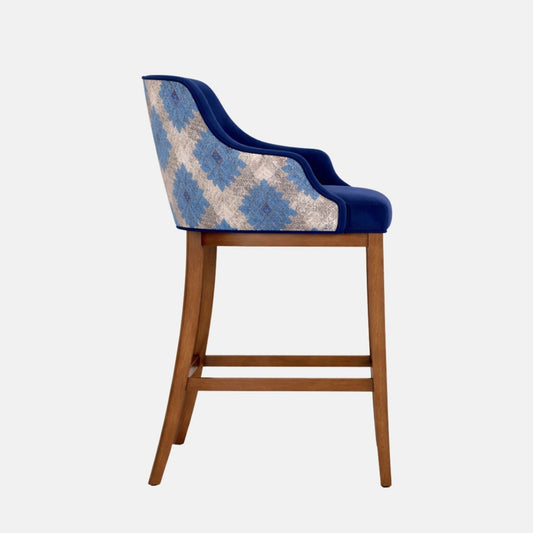 Front angled view of a bar-height stool with a navy upholstered seat and wraparound back/arms, patterned blue-and-cream fabric on the outer back panel, and a medium-brown wooden frame with four legs and a square footrest stretcher, photographed on a white background.